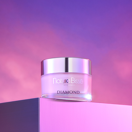 De Natura Bissé Diamond Body Cream pot gepresenteerd in een artistieke setting met een sculpturale zwarte vorm en een ruw kristal op een beige ondergrond.