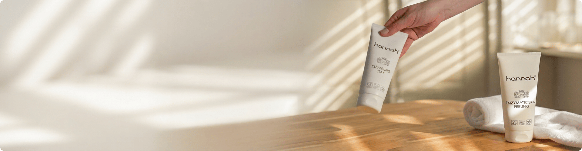 Een brede banner met een houten tafel in warm zonlicht waarop een hand een tube hannah Cleansing Clay vasthoudt naast een tube hannah Enzymatic Skin Peeling op een witte handdoek.