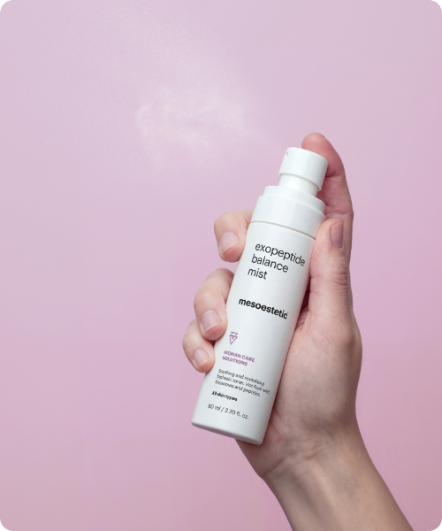 Een hand houdt een flacon met mesoestetic exopeptide balance mist vast tegen een roze achtergrond terwijl er een fijne nevel uit de spraykop komt