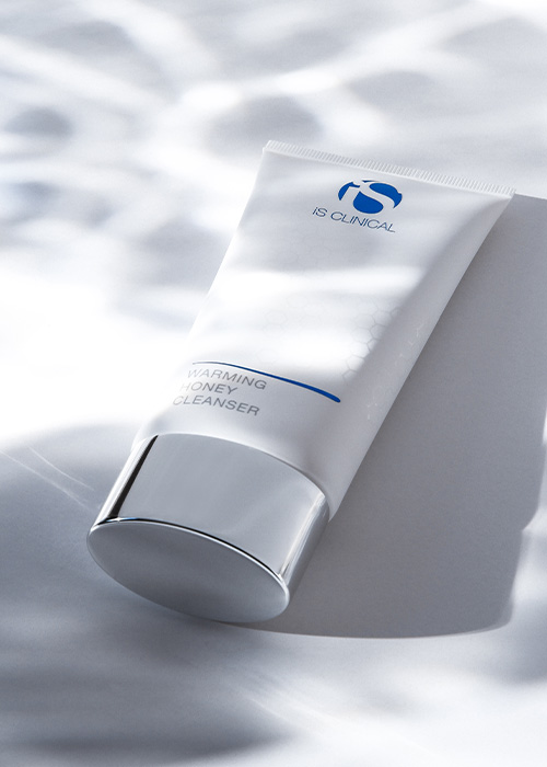 Een tube iS CLINICAL Warming Honey Cleanser op een witte ondergrond met artistieke schaduweffecten.
