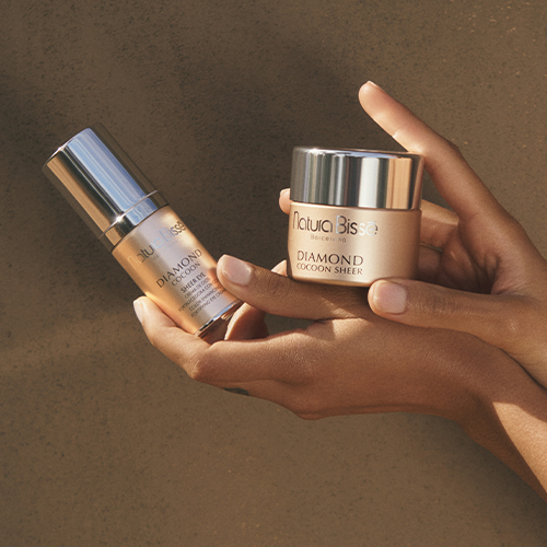 Iemand houdt de Diamond Cocoon Sheer Cream pot en de Sheer Eye dispenser vast tegen een warme, neutrale achtergrond.