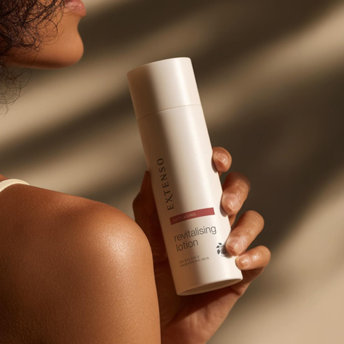 Een vrouw die de Extenso anti aging revitalising lotion fles vasthoudt bij haar schouder in warm zonlicht
