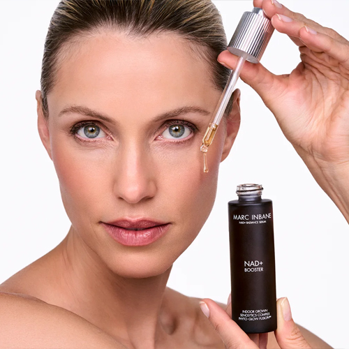 Close-up van een model dat een druppelaar met gouden serum bij haar gezicht houdt en een flesje Marc Inbane NAD+ Booster toont