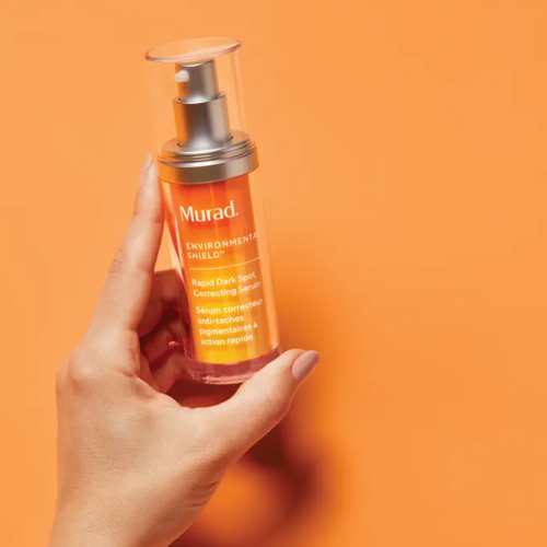 Hand die een flacon Murad Rapid Dark Spot Correcting Serum vasthoudt voor een oranje achtergrond