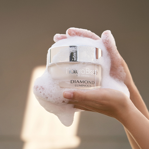 Twee handen die een pot Natura Bissé Diamond Luminous vasthouden, volledig bedekt met rijk wit reinigingsschuim.