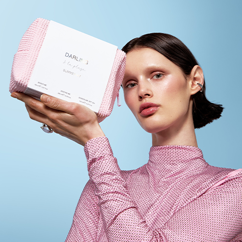 Een model met donker haar, felle make up en een roze met steentjes ingelegde top houdt de Darling A la plage Summer Kit toilettas omhoog tegen een lichtblauwe achtergrond.