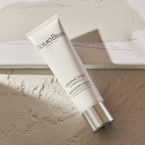 Een witte tube Natura Bissé Diamond Luminous Rich Luxury Cleanse op een artistieke beige achtergrond met verfstreken.