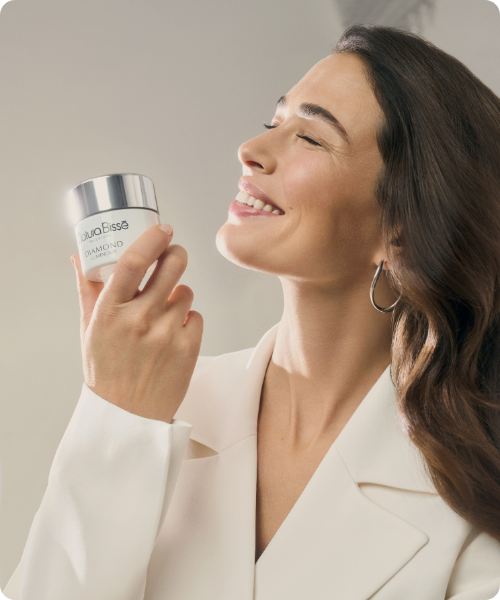 Een glimlachende vrouw in een wit jasje die een pot Natura Bissé Diamond Luminous Cream vasthoudt en ruikt aan het product.