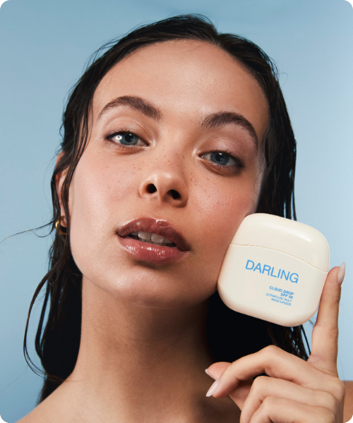 Een close-up van een model met een gehydrateerde huid die een compacte, witte flacon van DARLING Cloud Drop SPF 50 Ultrafluid Daily Moisturizer tegen haar wang houdt.