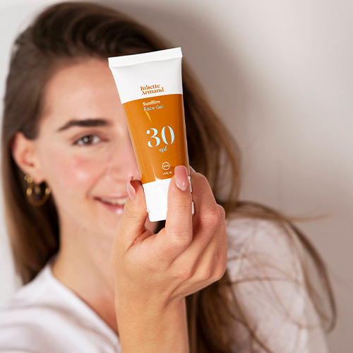 Een lachende vrouw houdt een tube Juliette Armand Sunfilm Face Gel SPF30 dicht bij de camera. De tube is wit met een opvallend oranje vlak en de tekst '30 SPF'. De achtergrond is licht en wazig.