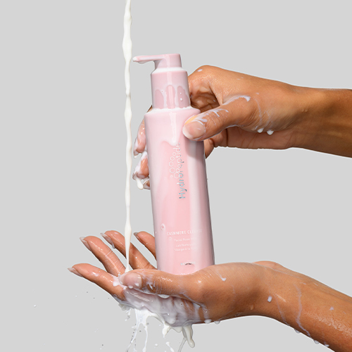 Handen houden een roze fles HydroPeptide Cashmere Cleanser vast terwijl er reinigingsmelk overheen stroomt tegen een grijze achtergrond