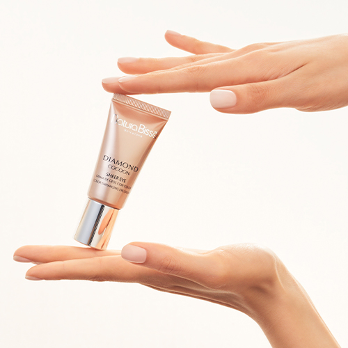 Twee paar handen met zachtroze nagellak houden de roze tube Natura Bissé gekleurde oogcrème vast op een zachte, crèmekleurige achtergrond.