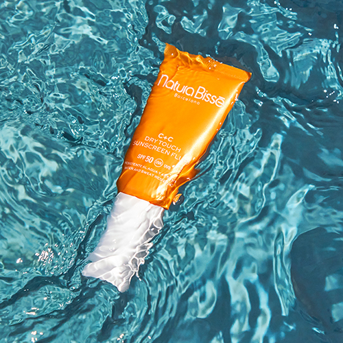 Een oranje tube Natura Bissé SPF 50 zonnebrandvloeistof drijvend in helderblauw, rimpelend zwembadwater, wat de waterbestendigheid benadrukt.