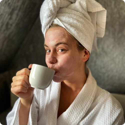Vrouw in een witte badjas en een handdoek om haar haar drinkt uit een mok terwijl ze een gezichtsmasker draagt.