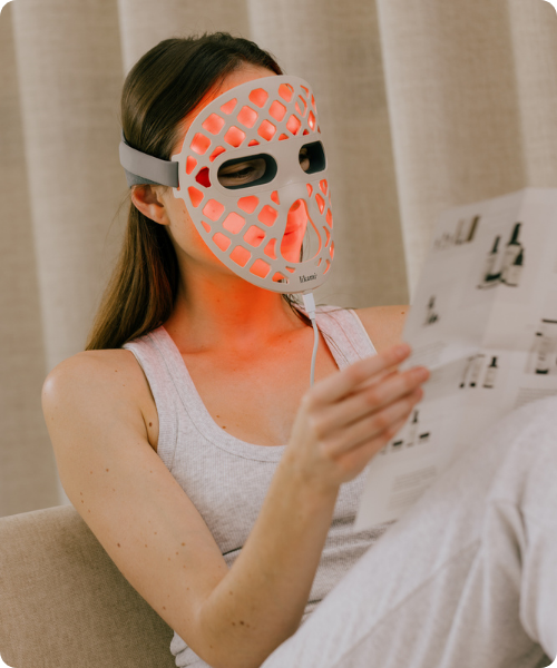 Een jonge vrouw met donker haar draagt een wit Likami LED gezichtsmasker dat rood oplicht terwijl ze in een brochure leest in een stoel