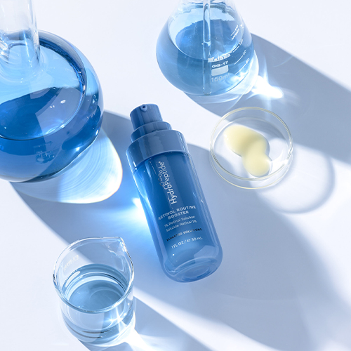 De blauwe flacon van de HydroPeptide Retinol Routine Booster omringd door laboratoriumglaswerk met blauwe vloeistof en een horlogeglas met de substantie.