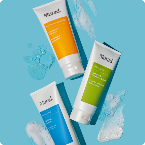 Drie tubes Murad cleansers – Essential-C Cleanser, Renewing Cleansing Cream en Clarifying Cleanser – plat neergelegd op een blauwe achtergrond met schuim- en geltexturen om de tubes heen.