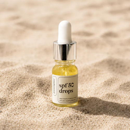 Het glazen pipetflesje met de gele Prescription SPF 50 drops staat rechtop in het fijne strandzand tijdens een zonnige dag.