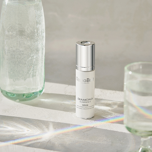 Een fles Natura Bissé Diamond Luminous Serum staat centraal op een stenen platform, tussen een glazen waterkaraf en een gevuld waterglas, met regenboogkleurige lichtstralen die over de scène schijnen.