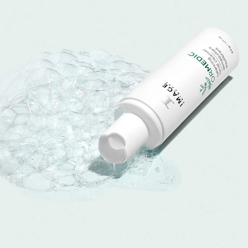 De witte tube van de ORMEDIC Balancing Facial Cleanser liggend naast een golvende swatch van de transparante reinigingsgel.