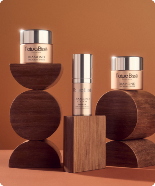 Drie luxe huidverzorgingsproducten uit de Diamond Cocoon lijn van Natura Bissé, gepresenteerd op een artistieke stapel van geometrische houten vormen tegen een warme, bruine achtergrond.