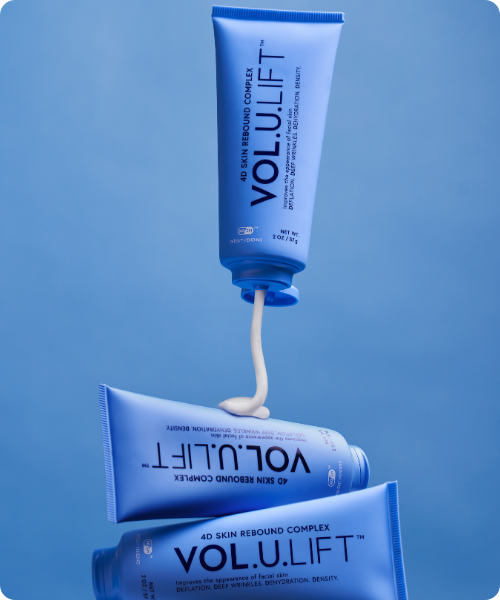 Een artistieke weergave van drie blauwe IMAGE Skincare VOL.U.LIFT tubes waarbij de bovenste tube een witte, romige substantie op de onderste tubes aanbrengt tegen een blauwe achtergrond.