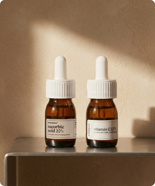 Twee bruine glazen flesjes met witte pipetdop en labels van Prescription op een metalen plank tegen een beige achtergrond, waarbij het linker flesje ascorbic acid 20% en het rechter flesje vitamin c 15% vermeldt.