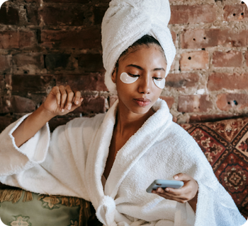 Een jonge vrouw in een witte badjas met een handdoek om haar haar en oogmaskers op, kijkt geconcentreerd op haar smartphone tegen een achtergrond van een bakstenen muur.
