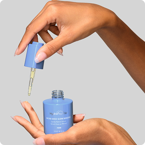 Een hand houdt een blauw flesje HydroPeptide Micro-Dose Glow Booster vast terwijl de andere hand het glazen pipetje boven het flacon tilt.