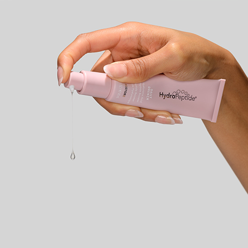 Handen die een roze tube HydroPeptide Daily Drench vasthouden waar een druppel vloeistof uitkomt tegen een grijze achtergrond