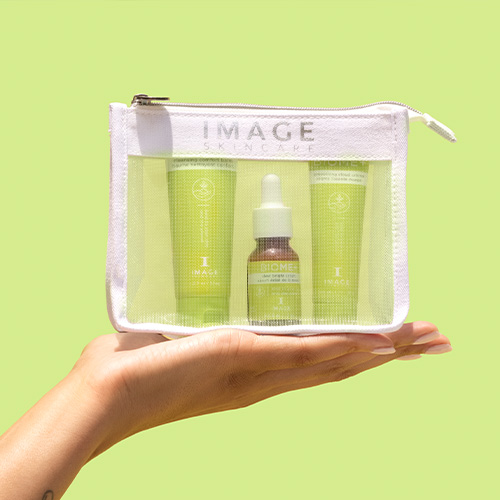 Een hand die de transparante IMAGE Skincare BIOME+ Discovery Kit vasthoudt tegen een lichtgroene achtergrond, met de reinigingsbalsem, het serum en de moisturizer zichtbaar in het tasje.