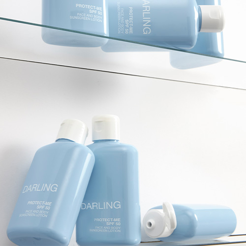 Meerdere blauwe flacons van DARLING PROTECT-ME SPF 50 op een glazen plank, waarvan één flacon geopend is.