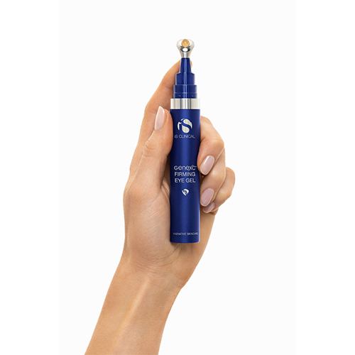 Een hand met verzorgde nagels houdt de blauwe huls van de iS Clinical Genexc Firming Eye Gel verticaal vast tegen een witte achtergrond