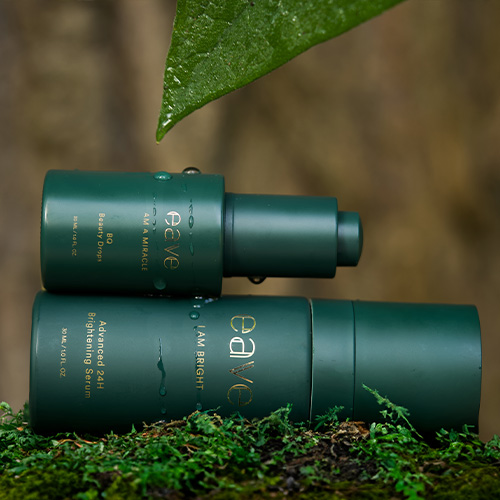 Twee donkergroene EAVE flacons, waaronder het I AM BRIGHT serum, liggend op groen mos met waterdruppels in een natuurlijke omgeving.
