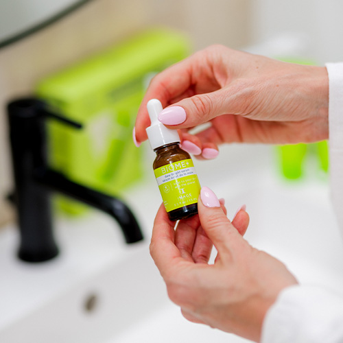 Detailopname van een vrouw met roze nagels die het glazen pipetflesje van het IMAGE Skincare BIOME+ Dew Bright Serum vasthoudt in een badkamer.