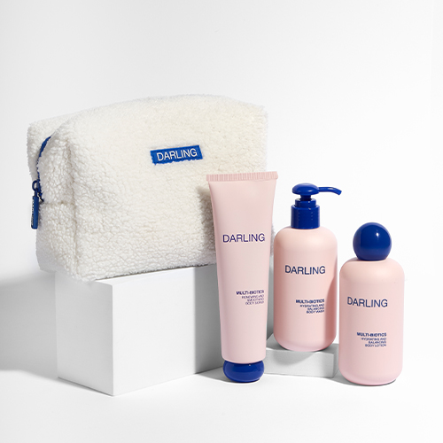 Een productfoto op een witte achtergrond van de Darling Multi Biotics set. De witte pluizige toilettas en de drie lichtroze lichaamsproducten staan op witte, vierkante displays.