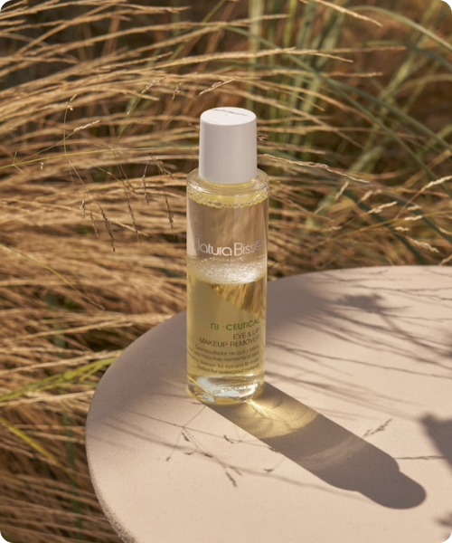 Een transparante flacon van de Natura Bissé NB-Ceutical Eye & Lip Makeup Remover staat op een ronde, beige tafel in de zon, met een achtergrond van wuivend, droog gras. De afbeelding heeft afgeronde hoeken.