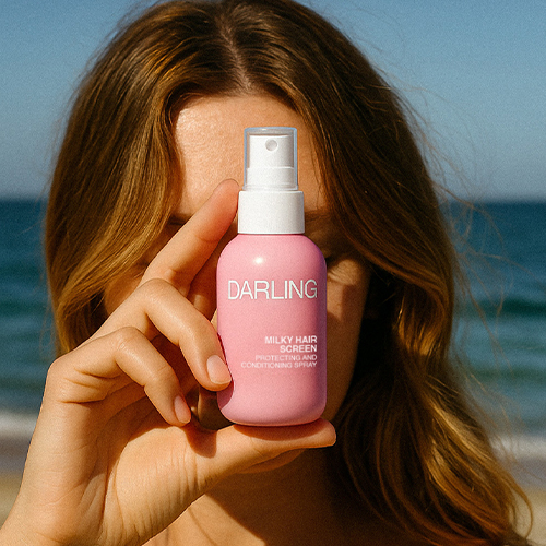 Een model houdt de travelsize Darling Milky Hair Screen spray voor haar gezicht met de zee op de achtergrond.