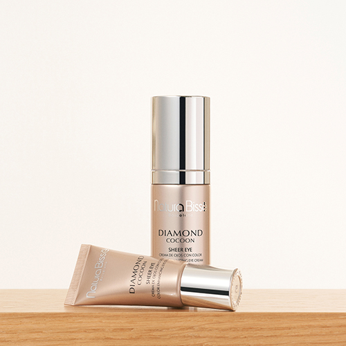 Een staande fles en een liggende tube van Natura Bissé gekleurde oogcrème op een licht houten plank tegen een crèmekleurige achtergrond.