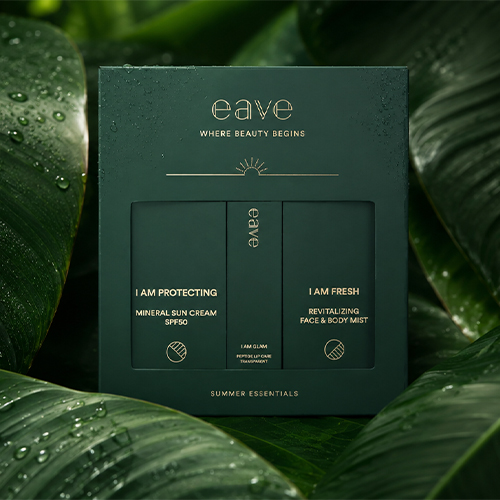 Een sfeerbeeld van de gesloten EAVE SUMMER ESSENTIALS BOX tussen grote, donkergroene, glanzende bladeren met waterdruppels.
