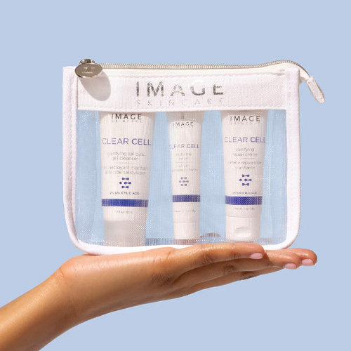 Een hand die de doorzichtige IMAGE Skincare tas vasthoudt met daarin drie CLEAR CELL blemish control producten tegen een lichtblauwe achtergrond.