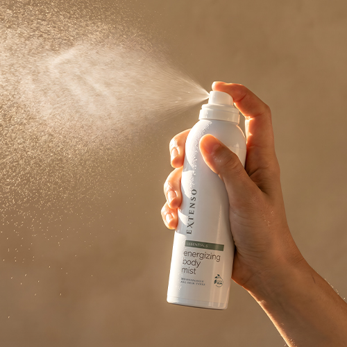 Een hand die de Extenso Energizing Body Mist sprayfles vasthoudt en een fijne nevel in de lucht spuit met een warme beige achtergrond