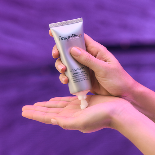 Close-up van handen die het witte Diamond Extreme Mask uit de zilveren tube knijpen tegen een sfeervolle paarse achtergrond.