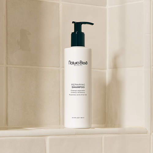 De witte fles Natura Bissé Repairing Shampoo staat op een betegelde planchet in een lichte, moderne badkamer.