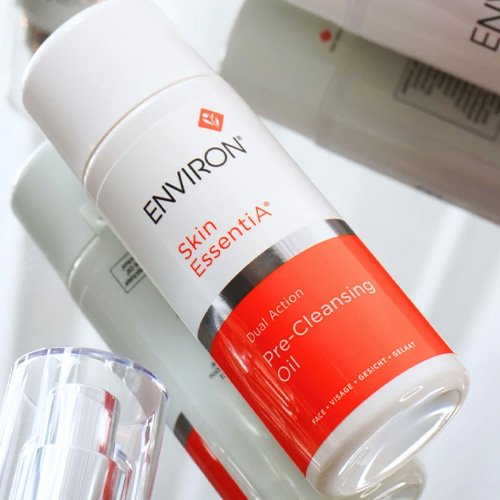 Een close-up van de Environ Skin EssentiA Dual Action Pre-Cleansing Oil flacon, liggend op een spiegelende ondergrond waarin andere Environ verpakkingen reflecteren.