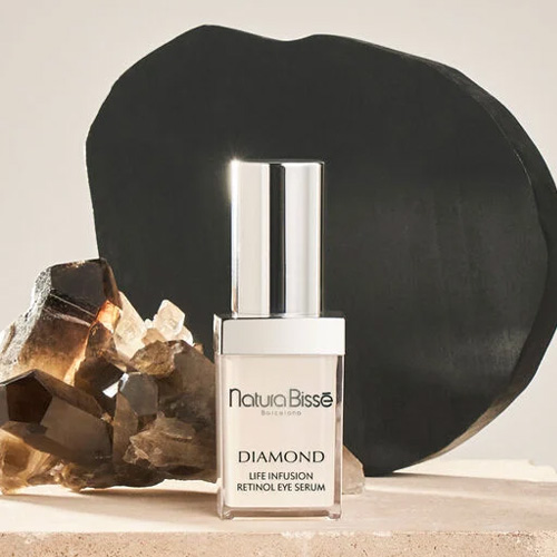 Een luxe glazen flacon van Natura Bissé Diamond Life Infusion Retinol Eye Serum voor een beige achtergrond, geflankeerd door een donker rookkwarts kristal en een zwarte organische vorm.