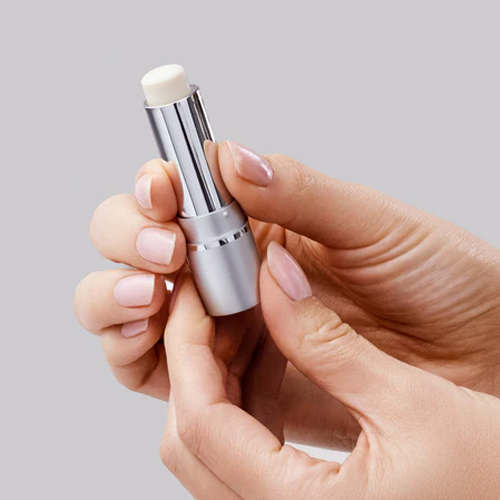 Handen die een geopende zilveren huls van iS Clinical Lip Protect SPF 35 lippenbalsem vasthouden tegen een grijze achtergrond