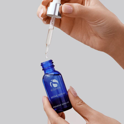 Handen die een blauw glazen pipetflesje van iS Clinical Active Serum vasthouden terwijl er een druppel uit de pipet valt tegen een grijze achtergrond