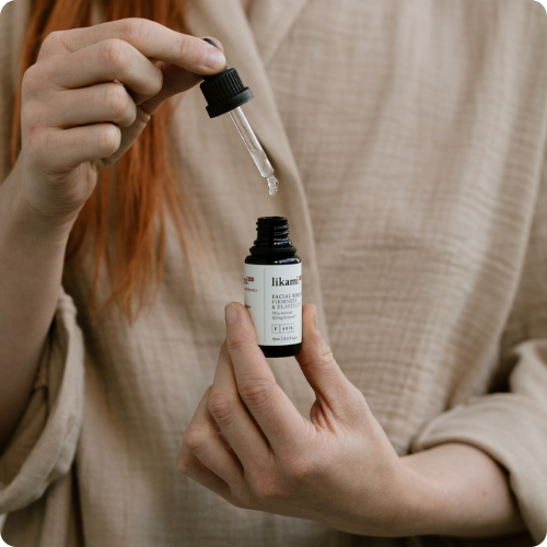 Een close-up van handen die een glazen flesje Likami Firmness & Elasticity serum vasthouden, terwijl er met een pipet een druppel wordt gedoseerd. De afbeelding heeft afgeronde hoeken.