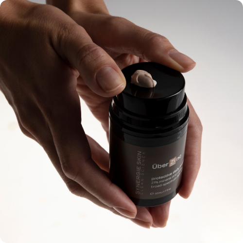 Twee handen die een pompflacon Synergie Skin ÜberZinc vasthouden met een toefje minerale crème op het pompje